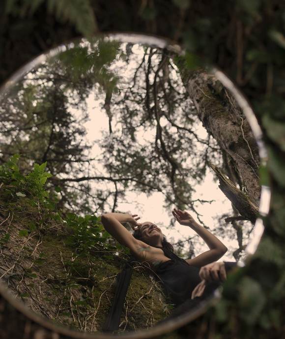 Ana, smiling in a forest, reflected in a circular mirror on mossy ground.